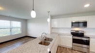 This single-floor space offers a clean, contemporary design with an open-concept layout that seamlessly connects the kitchen and living area. The kitchen is a standout feature, showcasing crisp white cabinetry, light granite countertops, and a full suite of stainless steel appliances including a refrigerator, electric range, and over-the-range microwave. A peninsula with a double undermount sink and elegant pendant lighting serves as a natural divider between the kitchen and the spacious living area, adding both functionality and visual appeal. Wood-look plank flooring flows consistently throughout the entire space, complemented by dark baseboards that provide a sharp contrast against the bright white walls. The living area is generously sized and benefits from multiple windows that invite an abundance of natural light and offer expansive views of the surrounding landscape. A dedicated bathroom features matching wood-look flooring, a vanity, and a towel bar, maintaining the cohesive design language found throughout the unit. A utility hallway provides access to the electrical panel and additional doorways, ensuring practical functionality. Recessed lighting and flush-mount ceiling fixtures provide ample illumination throughout, creating a bright and welcoming ambiance across every area of this thoughtfully designed space.