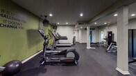 Located at 1025 Avent Hill in Raleigh, NC, this dedicated fitness center is a fully outfitted, single-floor facility designed to support a comprehensive workout experience. The space is defined by its bold aesthetic — vibrant lime-green accent walls contrast sharply with neutral gray tones on the ceiling and flooring, creating an energizing atmosphere reinforced by motivational wall decals, including the phrase "One More Rep."

The flooring throughout is durable rubber gym matting, ideal for both cardio and strength training areas. Recessed lighting provides consistent, bright illumination across the entire space, complemented by a ceiling fan that aids in air circulation during intense workouts.

The cardio section is well-stocked with multiple treadmills and elliptical machines, each positioned with wall-mounted flat-screen televisions nearby for entertainment. The strength training area includes a cable machine, adjustable weight benches, free weights, medicine balls, and a multi-functional rack system. Full-length wall mirrors along one side visually expand the space and allow users to monitor their form.

Structural support columns are integrated neatly into the layout, and an additional room accessible via an interior door appears to offer supplemental space. A fire extinguisher is also visibly mounted, ensuring safety compliance. Overall, this fitness center delivers a functional, motivating, and well-maintained environment for residents or members.