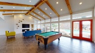 This single-floor community clubhouse blends mid-century modern design with open, airy architecture. The main gathering area commands attention with soaring vaulted ceilings adorned with exposed natural wood beams and recessed lighting, creating a bright and welcoming atmosphere. Large floor-to-ceiling windows line an entire wall, flooding the space with natural light and offering unobstructed views of the outdoor pool and surrounding landscape. A bold white textured brick accent wall serves as the room's focal point, featuring built-in shelving, a wall-mounted flat-screen television, and a fireplace below — all framed by mid-century inspired seating in vibrant yellow, blue, and orange upholstery set atop large-format dark tile flooring. A full-size billiards table anchors the game area, positioned to take advantage of the panoramic pool views through the glazed wall, with direct access to the outdoor area via a red-framed door. The adjacent kitchenette is well-appointed with white cabinetry, butcher block countertops, teal subway tile backsplash, and a dining area illuminated by a sculptural brass chandelier. Throughout, the space maintains a cohesive mid-century modern aesthetic with bold color accents, decorative sunburst wall art, and an open layout ideal for social gatherings.