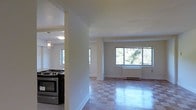 This single-floor space features an open-concept layout that seamlessly connects the kitchen and living areas. The kitchen is equipped with a stainless steel range and accented by elegant pendant lighting, adding a modern touch to the space. A structural column subtly defines the boundary between the kitchen and the living area without interrupting the overall sense of openness.

The living area is bright and airy, benefiting from a large multi-pane window that frames views of lush, mature trees outside, allowing abundant natural light to fill the room. A baseboard heating unit runs along the exterior wall, providing functional climate control while maintaining a clean aesthetic.

The flooring throughout features a checkered tile pattern that adds visual interest and reflects the natural light streaming in from the windows. The walls are finished in a soft, neutral tone, contributing to the clean and inviting atmosphere of the space.

Overall, the layout is practical and well-lit, with a calm, neutral palette that provides a versatile foundation for a variety of interior styles. The connection to greenery through the large windows enhances the sense of comfort and tranquility throughout.