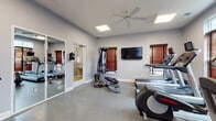 This fitness center is a thoughtfully designed, single-floor workout facility offering a comprehensive range of exercise equipment and amenities. The space is finished with durable speckled flooring throughout, providing a practical and easy-to-maintain surface suited for high-traffic athletic use. Neutral gray walls and white trim create a clean, modern aesthetic that keeps the environment feeling open and energizing.

The main workout area is well-equipped with multiple treadmills, elliptical machines, a stationary bike, a multi-function cable strength training machine, and a weight bench, catering to both cardiovascular and strength training needs. A wall-mounted flat-screen television is centrally positioned for entertainment during workouts, while a ceiling fan ensures comfortable air circulation throughout the space.

Natural light filters in through several windows fitted with rich wood-toned blinds, adding warmth to the otherwise contemporary interior. Full-length mirrored panels along one wall visually expand the space and allow users to monitor their form during exercise. A connected restroom is conveniently accessible directly from the gym floor. A separate entry door with an emergency exit sign ensures clear and safe access points. The overall ambiance is functional, bright, and well-appointed, making it a practical and inviting fitness destination.