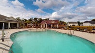 Located at 75 Siskin Cir in Hempstead, NC, this single-story community amenity center offers a well-appointed outdoor and indoor experience designed for relaxation and recreation. The centerpiece of the property is a generously sized, freeform resort-style swimming pool with a gradual entry, crystal-clear turquoise water, and a surrounding concrete pool deck lined with lounge chairs—ideal for sunbathing and socializing. Tall palm trees and carefully maintained landscaping with mulched planting beds provide a coastal aesthetic throughout the grounds, enhancing the welcoming atmosphere. The pool house structure is clad in light gray horizontal lap siding with crisp white trim and topped with a distinctive dark red metal roof, giving it a refined yet relaxed coastal character. A covered front porch features a ceiling fan with a pendant-style fixture, decorative area rugs, and comfortable seating, creating an inviting transition between the indoors and the pool area. An outdoor-mounted television on the building's exterior adds to the entertainment options available on the patio. Signage on the building indicates a dedicated fitness center accessible from the porch. The surrounding community includes residential buildings visible in the background, set within a landscaped neighborhood that complements the amenity center's coastal design theme.