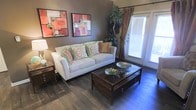 This single-floor space showcases a thoughtfully designed living area that balances comfort and style. The living room features rich, dark hardwood-style flooring that anchors the space and complements the warm taupe-toned walls. A white sofa dressed with colorful accent pillows in coral, green, and gold hues serves as the focal point, paired with a glass-topped wooden coffee table that adds an elegant touch. Two bold abstract artworks in coral and pink tones adorn the wall, adding a vibrant artistic element to the room. A wooden side table with a classic lamp provides warm ambient lighting. Natural light filters into the space through a large window dressed with decorative drapes in rich earth tones, while a glass door allows additional brightness and potential outdoor access. A tall indoor plant near the window introduces a refreshing natural element. The overall ambiance of the space is warm and welcoming, with a cohesive color palette of earthy neutrals accented by pops of coral and green, creating an inviting atmosphere suited for everyday living.