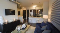 This single-floor space showcases an open-concept design that integrates the living area and kitchen into one cohesive, functional layout. The living area is anchored by a plush navy blue sofa, complemented by a sleek coffee table styled with coastal-inspired decorative accents. Dark hardwood flooring runs throughout, providing a rich contrast to the light-colored walls. A wall-mounted flat-screen television and a low-profile media console add to the room's modern aesthetic.

The kitchen features dark cabinetry with glass-front upper panels, a light stone countertop, and a peninsula that doubles as a breakfast bar with white bar stools. Pendant lighting above the peninsula adds warm, focused illumination, while stainless steel appliances, including a French door refrigerator, complete the contemporary kitchen setup. A tile backsplash adds texture and visual interest behind the range area.

An industrial character is introduced through exposed ductwork along the ceiling near the entryway, adding an urban loft-inspired element. Track lighting and recessed fixtures provide layered lighting throughout the space. The overall ambiance is modern and well-appointed, blending comfort with clean, contemporary design.