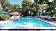 This outdoor pool area offers a resort-like retreat set beneath a canopy of tall, mature trees that provide natural shade and a serene, park-like atmosphere. The generously sized rectangular pool features vivid blue tile detailing along its edges and steps, with clear, well-maintained water that reflects the surrounding greenery and open sky. Depth markers are visible along the pool's edge, indicating a thoughtfully designed swimming environment suitable for a range of swimmers. The expansive pool deck is finished in neutral tiling and furnished with multiple lounge chairs and seating arrangements, inviting relaxation before or after a swim. A charming single-story cabana or pool house structure sits adjacent to the pool, featuring a covered porch with white railings, providing a shaded gathering space and likely housing amenities such as restrooms or equipment storage. A white perimeter fence encloses the area, ensuring privacy and safety. Blue patio umbrellas add a pop of color to one side of the deck. Together, these elements create a welcoming, well-appointed outdoor recreational space ideal for leisure and relaxation.
