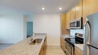 This single-floor space offers a clean, contemporary design with a strong focus on functionality and modern finishes. The kitchen serves as a central highlight, showcasing warm honey-toned wood cabinetry that extends from floor to ceiling, providing generous storage capacity. Granite countertops in a speckled neutral palette run throughout, offering ample workspace and a polished aesthetic. A stainless steel undermount sink is positioned on a peninsula-style counter, which adds both workspace and a natural division between the kitchen and adjacent areas. Stainless steel appliances, including an electric range with a smooth cooktop and an over-the-range microwave, complete the kitchen's professional feel. Light hardwood flooring runs throughout the space, contributing to an open and airy atmosphere. Recessed lighting in the white ceiling ensures the kitchen is well-lit and inviting. Crisp white walls keep the overall palette bright and neutral, complementing the warm wood tones. A connected room visible through a doorway hints at additional living areas, rounding out this thoughtfully designed, move-in-ready space.