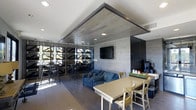 This single-floor space showcases a thoughtfully designed interior that blends modern aesthetics with functional versatility. A striking suspended ceiling panel clad in wood-look material serves as a defining architectural feature, anchoring the central gathering area and adding warmth against the cool, light-toned stone-textured accent walls.

The space is organized into multiple distinct zones. A dining area near the foreground features a light-colored table paired with wooden chairs, suitable for casual meals or collaborative work. Adjacent to it, a comfortable lounge area offers a blue sofa and armchair arrangement, creating an inviting setting for relaxed conversation. A coffee and beverage station is positioned along the far wall, equipped with machines and cup supplies for easy self-service.

Toward the rear, a bar-height seating area with tall stools and industrial-style metal shelving provides an additional social zone, bathed in natural light from large windows that line the perimeter. The dark hardwood-style flooring unifies all areas cohesively. Recessed lighting supplements the natural light, maintaining a bright and welcoming ambiance throughout.