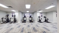 Located at 425 W 5th St in uptown Charlotte, NC, this fitness center offers a well-appointed, single-floor layout designed to support a full range of workout routines. The space features a clean, modern aesthetic with light gray walls, a drop tile ceiling with recessed panel lighting, and stylish blue-accented carpet tiles that run throughout.

The cardio zone includes multiple treadmills and stationary cycling bikes, each equipped with individual digital screens, positioned near arched and rectangular windows that allow natural light to filter in and provide views of the surrounding urban environment. A dedicated strength training area houses a multi-station cable machine, a weight bench, and a rack of dumbbells, complemented by a wall-mounted flat-screen TV and a mirrored wall that enhances the sense of space.

A functional training section is stocked with stability balls, medicine balls, resistance bands, foam rollers, and a BOSU trainer, all neatly organized on a freestanding rack alongside another wall-mounted display screen. Additional strength equipment, including a leg press and cable attachment stations, occupies a separate open zone.

The facility's spacious, elongated floor plan allows for comfortable movement between zones. Structural columns are integrated throughout without impeding the flow of the space. The overall ambiance is bright, clean, and professionally outfitted, making it a comprehensive fitness destination in the heart of Charlotte.