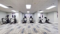 Located at 425 W 5th St in uptown Charlotte, NC, this fitness center offers a well-appointed, single-floor layout designed to support a full range of workout routines. The space features a clean, modern aesthetic with light gray walls, a drop tile ceiling with recessed panel lighting, and stylish blue-accented carpet tiles that run throughout.

The cardio zone includes multiple treadmills and stationary cycling bikes, each equipped with individual digital screens, positioned near arched and rectangular windows that allow natural light to filter in and provide views of the surrounding urban environment. A dedicated strength training area houses a multi-station cable machine, a weight bench, and a rack of dumbbells, complemented by a wall-mounted flat-screen TV and a mirrored wall that enhances the sense of space.

A functional training section is stocked with stability balls, medicine balls, resistance bands, foam rollers, and a BOSU trainer, all neatly organized on a freestanding rack alongside another wall-mounted display screen. Additional strength equipment, including a leg press and cable attachment stations, occupies a separate open zone.

The facility's spacious, elongated floor plan allows for comfortable movement between zones. Structural columns are integrated throughout without impeding the flow of the space. The overall ambiance is bright, clean, and professionally outfitted, making it a comprehensive fitness destination in the heart of Charlotte.