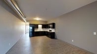 This single-floor space presents an open-concept layout that seamlessly integrates the living area with a fully equipped kitchen. The kitchen is outfitted with deep espresso-toned cabinetry that extends along two walls in an L-shaped configuration, creating an efficient and visually striking contrast against the light-colored walls and white tile backsplash. Black appliances, including a full-size refrigerator and range, complement the dark cabinetry and contribute to a cohesive, contemporary aesthetic. A stainless steel sink is positioned beneath the cabinetry, offering functional workspace throughout. Light hardwood-style flooring spans the entire space, adding warmth and continuity between the kitchen and the open living area. The ceilings are smooth and neutral, contributing to a clean, understated ambiance. Exposed ductwork is visible along one wall, lending a subtle industrial character to the space. A doorway visible at the far end suggests access to additional rooms or a balcony, offering potential for natural light. Overall, the space balances modern design elements with practical functionality in a spacious, open arrangement.