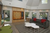 This single-floor space makes a strong architectural impression with its dramatically tall ceilings and an open, flowing layout that encourages a sense of airiness and volume. Large windows frame lush greenery outside, flooding the interior with natural light and creating a seamless visual connection to the outdoors.

The main living area is anchored by a substantial dark sofa accented with bold red cushions, positioned atop a graphic black-and-white area rug. Flanking this central seating zone are woven rattan armchairs and a warm leather bench, contributing a rich layering of textures and tones. A pair of sleek white coffee tables grounds the arrangement, while sculptural wall-mounted lighting adds focused ambiance.

Along one wall, a full-height built-in shelving unit crafted from light natural wood provides generous display and storage space. The shelves are thoughtfully styled with artwork, books, and decorative objects. Adjacent to this, a cluster of green cushioned accent chairs on wire-frame bases introduces a playful mid-century modern touch.

The flooring throughout is wide-plank light wood, lending warmth and continuity across the space. Structural white columns punctuate the room, reinforcing the architectural character while defining distinct seating zones. Overall, the space balances contemporary design sensibility with curated, inviting furnishings.