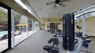 This fitness center offers a thoughtfully designed workout environment that balances functionality with an inviting atmosphere. The space is defined by expansive floor-to-ceiling windows along one side, flooding the interior with abundant natural light and providing clear views of an outdoor patio area complete with covered seating and lush greenery. The open layout accommodates a well-rounded selection of exercise equipment, including a row of cardio machines such as treadmills and ellipticals, as well as a Life Fitness multi-station strength training unit and a free-weight bench. The carpeted flooring provides a comfortable surface throughout the workout area. The ceiling features multiple ceiling fans evenly spaced along the length of the room, ensuring consistent air circulation during exercise. Recessed and pendant lighting supplements the natural light for evening or low-light use. The neutral, warm-toned walls contribute to a clean and focused environment. At the far end of the space, a built-in unit appears to house additional amenities. Overall, this fitness center delivers a bright, airy, and well-appointed setting suitable for a comprehensive workout routine.