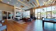 This upscale resident lounge is a thoughtfully designed amenity space spread across a single floor, offering a variety of distinct zones for relaxation, socializing, and entertainment. The main lounge area commands attention with soaring ceilings, a coffered ceiling detail accented in gold, and a dramatic sculptural chandelier. A sleek linear fireplace set into a floor-to-ceiling stone accent wall serves as a striking focal point, flanked by curated seating arrangements on rich hardwood flooring. Expansive floor-to-ceiling windows flood the interior with natural light and provide sweeping views of the surrounding outdoor amenity areas. A dedicated café and dining area features polished concrete flooring, bar-height seating along a marble countertop, and contemporary pendant lighting, creating an inviting space for casual gathering. Adjacent to this is a private dining room appointed with a live-edge wood table, upholstered chairs, and curated artwork beneath a sculptural light fixture. The demonstration kitchen is fully equipped with professional-grade stainless steel appliances, a large quartz island, and bar seating. The entertainment lounge rounds out the space with a billiards table, a built-in granite wet bar with wine refrigeration, and stylish decorative mirrors, making it an ideal venue for social events.