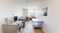 Located at 318–352 Patricia Ave in Kitchener, Ontario, this shared laundry facility offers a clean, well-organized space designed for practical, everyday use. The room is finished with light wood laminate flooring and bright white walls, creating a tidy and welcoming environment. Overhead fluorescent lighting ensures the space is well-lit throughout, supplemented by a window that brings in natural light and offers a view of the surrounding greenery.

The laundry room is equipped with multiple front-loading washing machines and dryers finished in a sleek silver tone, arranged efficiently along the walls to maximize usable floor space. A stainless steel folding and sorting table runs along one side of the room, providing ample surface area for handling laundry with ease. A utility sink is also present, adding further functionality to the space.

A digital payment terminal is conveniently mounted near the machines, offering a modern, card-free payment solution for users. Instructional and informational signage is posted on the walls, ensuring straightforward operation. The room is accessed through a solid entry door with secure hardware. The overall layout is thoughtfully arranged to accommodate multiple users comfortably, making this laundry room a highly functional shared amenity within the building.