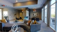 This refined single-floor space exudes contemporary sophistication through its thoughtfully curated design and premium finishes. At the heart of the room stands a dramatic floor-to-ceiling stone fireplace with a sleek, multi-sided surround, serving as a stunning architectural focal point that anchors the entire space. The lounge area is furnished with a generously sized curved sectional sofa in a rich charcoal upholstery, accented by warm cognac leather cushions and an assortment of earth-toned throw pillows, creating an inviting and stylish atmosphere. Adjacent seating areas feature dark upholstered armchairs and sofas arranged to encourage conversation and relaxation. Warm ambient lighting is provided by statement drum pendant fixtures and recessed ceiling lights, casting a soft, welcoming glow throughout. A tray ceiling detail adds architectural depth overhead. Large floor-to-ceiling windows along one wall allow natural light to filter into the space, enhancing its open, airy feel. Framed wall art adds curated visual interest, while decorative sculptural accents on the coffee tables complete the polished, upscale aesthetic. The overall ambiance is one of modern luxury — warm, welcoming, and meticulously designed.