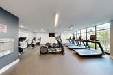 Health Hub is a thoughtfully designed fitness facility that spans a single, expansive floor, providing members with a comprehensive environment for all aspects of physical training. The space is organized into distinct zones, each purpose-built to support a specific type of workout.

The cardio area features a generous row of treadmills positioned alongside large floor-to-ceiling windows, flooding the space with natural light and offering views to the outdoors. Elliptical trainers and stationary cycling bikes round out the cardio offerings, all equipped with individual display screens. Linear LED lighting runs throughout the ceilings, providing bright, even illumination across all areas.

The strength training zone is well-appointed with a full dumbbell rack, weight benches, a cable machine, and a multi-functional power rack with TRX suspension equipment. A dedicated functional training area features a branded workout mat, resistance tools, and open floor space for dynamic movement exercises.

The flooring throughout is a durable, speckled rubber surface suited for high-traffic athletic use. Walls are finished in a clean white and deep navy-blue palette, contributing to a focused and energizing atmosphere. Mirrored walls and wall-mounted flat-screen televisions are strategically placed throughout. The entry area features black-framed glass doors, providing a sleek transition into the facility. Overall, the Health Hub delivers a modern, professional fitness experience.