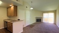 This single-floor residence is designed with an open-concept layout that creates a sense of spaciousness and ease of flow between living areas. The living room is a standout feature, boasting soaring vaulted ceilings that amplify the sense of volume and airiness throughout the space. A tiled fireplace with a clean, classic surround serves as the focal point of the living area, adding warmth and character. Plush carpeting covers the living room floor, providing comfort underfoot, while a sliding glass door leads to an outdoor area, allowing natural light to filter in and offering a seamless indoor-outdoor connection.

The kitchen is thoughtfully integrated into the open floor plan, separated from the living area by a raised breakfast bar finished with granite-style countertops. Warm wood cabinetry provides ample storage, and a pendant light fixture adds a decorative touch above the kitchen area. The kitchen also features a sink and functional layout suited for everyday use.

Two interior doors are visible, likely leading to additional rooms or storage areas. HVAC vents are positioned throughout, ensuring year-round comfort. Overall, the space presents a functional, well-designed layout with notable architectural details that enhance both its practicality and visual appeal.