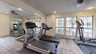 This fitness center offers a comprehensive workout environment spread across a single-level layout. The space is bright and welcoming, with large multi-pane windows lining the walls and allowing abundant natural light to fill the room. Recessed lighting and ceiling fans supplement the illumination and ensure a comfortable atmosphere during exercise.

The cardio zone is prominently featured and includes multiple Cybex treadmills and a selection of upright and recumbent stationary bikes, all positioned to take advantage of the outdoor views through the surrounding windows. Full-length mirrored walls amplify the sense of space and provide visual feedback during workouts.

A dedicated strength training area houses a multi-station cable and weight machine, along with additional resistance equipment and exercise mats for floor-based training. Rubber flooring in the weight zones transitions into patterned carpet tiles throughout the main area, providing both safety and comfort underfoot.

The space also features a textured ceiling, neutral warm-toned walls, and a set of French doors that open directly to an outdoor pool deck, seamlessly connecting indoor fitness with outdoor amenities. Safety features such as a fire extinguisher station are also present. The overall layout is functional and organized, providing users with a well-rounded facility suited for a full range of fitness activities.