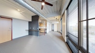 This single-floor space showcases a compelling blend of industrial and contemporary design. The soaring ceilings are defined by exposed galvanized ductwork and corrugated metal, lending the space a raw, urban character that is balanced by clean, modern finishes throughout. A large ceiling fan anchors the main living area, contributing both function and style.

The open-plan layout connects the living area seamlessly to the kitchen, which features light wood cabinetry, open shelving, and pendant lighting that creates a warm, inviting atmosphere. A built-in counter with a dark wood top and bold red metal legs serves as a striking room divider, providing a casual breakfast bar or workspace while maintaining visual flow between areas.

Floor-to-ceiling windows line one side of the space, fitted with horizontal blinds that allow for flexible light control. These expansive windows draw in generous amounts of natural light and offer a connection to the outdoors. The patterned carpet flooring adds texture underfoot, softening the industrial aesthetic of the upper elements. Track lighting and pendant fixtures are thoughtfully positioned throughout, ensuring the space is well-lit at all times. Overall, the space exudes a modern, loft-inspired ambiance that is both functional and visually dynamic.