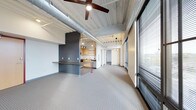This single-floor space showcases a compelling blend of industrial and contemporary design. The soaring ceilings are defined by exposed galvanized ductwork and corrugated metal, lending the space a raw, urban character that is balanced by clean, modern finishes throughout. A large ceiling fan anchors the main living area, contributing both function and style.

The open-plan layout connects the living area seamlessly to the kitchen, which features light wood cabinetry, open shelving, and pendant lighting that creates a warm, inviting atmosphere. A built-in counter with a dark wood top and bold red metal legs serves as a striking room divider, providing a casual breakfast bar or workspace while maintaining visual flow between areas.

Floor-to-ceiling windows line one side of the space, fitted with horizontal blinds that allow for flexible light control. These expansive windows draw in generous amounts of natural light and offer a connection to the outdoors. The patterned carpet flooring adds texture underfoot, softening the industrial aesthetic of the upper elements. Track lighting and pendant fixtures are thoughtfully positioned throughout, ensuring the space is well-lit at all times. Overall, the space exudes a modern, loft-inspired ambiance that is both functional and visually dynamic.