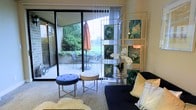 This single-floor residence offers a thoughtfully curated living experience that seamlessly blends indoor comfort with outdoor living. The main living area is bright and welcoming, anchored by large floor-to-ceiling sliding glass doors that open directly onto a private patio. The patio extends the living space outdoors, featuring seating and a patio umbrella, all framed by mature, verdant greenery that lends a serene, park-like atmosphere.

Inside, the living area is finished with plush carpeting and styled with an eclectic mix of contemporary furnishings, including a deep navy sofa accented with decorative pillows, round tufted ottomans with gold-toned frames, and a cozy faux fur area rug. A striking gold-framed display shelf showcases rich, jewel-toned abstract artwork in deep greens and teals, adding a bold artistic focal point to the room. Delicate white orchids and elegant accent lighting contribute to the refined, gallery-like ambiance.

Sheer vertical blinds filter natural light throughout the day, casting a soft glow across the space while maintaining privacy. The overall aesthetic is modern yet warm, combining artistic sensibility with functional design to create a residence that feels both sophisticated and livable.