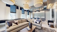 This thoughtfully designed single-floor space offers a generous, open-concept layout that seamlessly integrates a living room, dining area, and kitchen into one cohesive entertaining environment. Soaring ceilings and expansive floor-to-ceiling windows flood the interior with natural light while framing views of the surrounding exterior. The living area is anchored by oversized drum pendant lighting and features plush, neutral-toned seating complemented by navy blue accent pillows, layered area rugs over light wood-style flooring, and a wall-mounted flat-screen television. A striking starburst wall sculpture adds an artistic focal point alongside a built-in workspace with velvet blue accent chairs. The dining area includes a substantial dark-topped island with seating, illuminated by sleek pendant fixtures. The kitchenette is a standout feature, showcasing a bold geometric patterned tile backsplash, dark stone countertops, a matte black double-basin sink, and stainless steel appliances. A coffee and refreshment station is integrated into the space, making it ideal for communal gatherings. Large glass doors open directly to an outdoor patio, extending the living space and enhancing the bright, airy ambiance throughout. The overall aesthetic is modern and sophisticated, defined by a cohesive navy, grey, and gold palette.