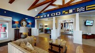 Located at 900 Old Fashioned Way in Newport, NC, this single-story community clubhouse delivers an impressive blend of nautical character and functional amenity spaces. The grand lounge anchors the facility with soaring vaulted ceilings supported by rich wood beam trusses, deep navy blue accent walls, and warm hardwood floors laid in a classic herringbone pattern. A fireplace, comfortable seating, and a built-in work area define this welcoming gathering space, while nautical signal flags displayed along the upper wall reinforce the coastal theme throughout. Built-in cabinetry with dark countertops and wood-paneled accents lines an adjacent room, providing additional flex space ideal for meetings or recreation. The fully equipped kitchen features white cabinetry, dark granite countertops, stainless steel appliances, and a large curved bar with seating — illuminated by pendant and track lighting with warm wood detailing above. A bright dining nook with large windows offers outdoor views and a wall-mounted TV. The fitness center is well-appointed across two connected rooms, housing treadmills, strength machines, and cardio equipment beneath a wood-paneled tray ceiling with a ceiling fan. A dedicated kids' room features whimsical underwater murals, natural light from multiple windows, and carpeted floors for play. Together, these spaces create a cohesive, resort-style community environment.