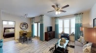 This single-floor space features an open-plan layout that seamlessly connects the living and dining areas, creating a cohesive and functional environment. The light-toned, wood-look laminate flooring runs throughout, adding warmth and visual continuity across the space.

The living area is anchored by a glass-top coffee table paired with a dark leather accent chair, while a flat-screen TV rests atop a rich, dark wood media console with glass-panel cabinet doors. A five-blade ceiling fan with an integrated light fixture is centrally positioned, providing both airflow and ambient lighting.

Large windows fitted with white horizontal blinds line multiple walls, allowing generous natural light to fill the room. Teal and white patterned curtains frame these windows, introducing a pop of color that ties the décor together.

The dining area, positioned toward one corner, accommodates a bar-height table surrounded by patterned yellow-green upholstered chairs, complemented by a decorative round wall mirror. A track lighting fixture overhead provides additional task lighting for the dining zone.

The textured ceiling and neutral wall tones create a clean, versatile backdrop throughout the space.