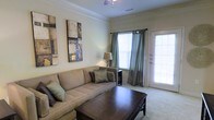 This single-floor space showcases a thoughtfully designed living room that serves as its centerpiece. The room is finished with classic crown moulding along the ceiling, adding a refined architectural touch, while a ceiling fan provides both function and a sense of vertical interest. Neutral, warm-toned walls create a calm and inviting ambiance throughout.

Natural light fills the space generously through a large window dressed with elegant sage green curtains, complemented by a glass-paneled door that provides direct access to the outdoors, further brightening the interior. The carpeted flooring adds warmth and comfort underfoot.

The living area is well-proportioned, accommodating a plush beige sofa adorned with coordinating decorative pillows and anchored by a dark wood coffee table. A side table with a classic lamp adds a soft, layered lighting option for the evenings. Abstract wall art in earthy tones provides a focal point and contributes to the space's contemporary yet approachable aesthetic.

Overall, the space offers a comfortable and polished atmosphere, combining practical features with tasteful design details suited for everyday living.