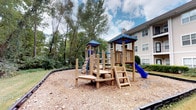 This residential community amenity space is thoughtfully designed to offer both comfort and functionality across a single level. The interior lounge area makes an immediate impression with its soaring ceilings, light wood-look flooring, and crisp white walls that create a bright, airy atmosphere. A grand fireplace serves as the focal point of the main living area, framed by an elegant white mantel and complemented by framed artwork above. The open layout seamlessly connects to a modern kitchen space outfitted with stainless steel appliances, a range hood, white cabinetry, and a kitchen island — ideal for community gatherings or events. A seating nook nearby features wingback chairs, a concrete coffee table, a large gold-framed floor mirror, and lush tropical greenery, adding a refined yet welcoming touch. Expansive windows flood the space with natural light and offer direct views of an outdoor pool area, where lounge seating can be seen. Outside, the community grounds include a well-equipped children's playground with a slide and climbing structures set on a wood chip safety surface, as well as a serene green space with a picnic table and charcoal grill, all surrounded by mature trees providing natural shade and privacy.
