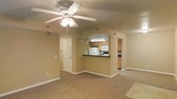 This single-floor residence features an open-concept layout that connects the living area to the kitchen through a convenient pass-through counter with a dark laminate surface, creating a sense of continuity and ease of interaction between spaces. The living area is finished with wall-to-wall carpet in a warm neutral tone, contributing to a cozy and practical atmosphere. A ceiling fan with a built-in light fixture is centrally mounted, providing both air circulation and ambient lighting. The kitchen is equipped with light oak cabinetry, a white refrigerator, a built-in microwave, and a range, offering ample functionality for everyday cooking. An additional cabinetry run is visible through a secondary kitchen opening, suggesting further storage and counter space. The walls throughout are painted in a soft beige tone, complementing the warm, neutral palette of the flooring and cabinetry. White six-panel doors and white baseboards add clean architectural detail throughout the space. Recessed accent lighting is visible along the walls, adding supplemental illumination options. Overall, the space presents a straightforward, well-maintained interior with practical features and a cohesive design aesthetic.