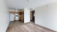This single-floor unit presents an open-concept layout that seamlessly connects the living area and kitchen, creating a functional and cohesive space. The living area features neutral-toned carpet that captures natural light streaming through windows, lending warmth to the room. A textured ceiling adds dimension overhead, while a ceiling fan in the kitchen area provides practical ventilation and air circulation.

The kitchen is efficiently designed with warm wood-toned cabinetry, a white electric range, a refrigerator, and under-cabinet space for storage. The kitchen floor transitions to a hard surface, contrasting with the carpeted living area and clearly delineating the two zones. A partial dividing wall separates the kitchen from the living space while maintaining an open feel.

A hallway visible from the main living area leads to additional rooms, including what appears to be a carpeted bedroom with a window allowing natural light. The HVAC vent integrated into the ceiling ensures comfortable climate control throughout the unit. Overall, the space presents a clean, straightforward layout suited for everyday living.