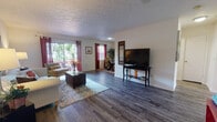 This single-floor home offers a thoughtfully laid-out living space with a warm, inviting atmosphere throughout. The expansive living area is anchored by rich, dark hardwood flooring that extends across the open floor plan. Large sliding glass doors and multiple windows allow natural light to pour in, offering views of the surrounding greenery. Vibrant curtains in deep red add a pop of color to the bright, white walls, while a textured ceiling adds architectural character.

The galley-style kitchen is efficiently designed with ample light wood cabinetry providing generous storage, a double sink, and full appliances including a dishwasher and refrigerator. The layout maximizes counter space and connects seamlessly to the rest of the home.

The bedrooms are carpeted for comfort and include closet space, with one bedroom featuring a walk-in closet. Neutral walls and textured ceilings continue throughout, creating a cohesive feel. A secondary room also provides dedicated dresser and storage space.

The bathroom is clean and functional, featuring a wide vanity with a large mirror, white cabinetry, and ample counter space. Coastal-themed artwork throughout the home ties the design together with a relaxed, breezy aesthetic, making this a comfortable and cheerful living space.