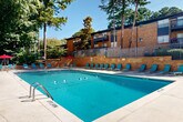 Located at 3901 Campbellton Rd SW in Atlanta, Georgia, this outdoor pool area serves as a central community amenity set against a backdrop of mature pine and deciduous trees. The generously sized rectangular pool features clearly marked depth zones, ranging from a shallow end to an 8-foot deep end, with lane lines dividing the water for structured use. A no-diving policy is posted, reinforcing the facility's safety standards.

Surrounding the pool is a well-maintained concrete deck lined with teal-colored lounge chairs, providing ample space for sunbathing and relaxation. On one side, a covered pergola area with red shade umbrellas offers a shaded gathering spot for residents. A two-story light-colored building with French doors sits directly adjacent to the pool, likely housing restrooms or a clubhouse facility, and provides easy access to and from the water.

The pool is enclosed by a metal security fence, with a warm brick retaining wall on one side that visually connects the area to the surrounding multi-story brick residential buildings featuring balconies. The lush, tree-lined setting creates a naturally shaded and private atmosphere, offering a refreshing outdoor retreat. The overall layout is thoughtfully designed to accommodate both active swimming and leisurely outdoor enjoyment throughout the warmer months.