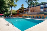 Located at 3901 Campbellton Rd SW in Atlanta, Georgia, this outdoor pool area serves as a central community amenity set against a backdrop of mature pine and deciduous trees. The generously sized rectangular pool features clearly marked depth zones, ranging from a shallow end to an 8-foot deep end, with lane lines dividing the water for structured use. A no-diving policy is posted, reinforcing the facility's safety standards.

Surrounding the pool is a well-maintained concrete deck lined with teal-colored lounge chairs, providing ample space for sunbathing and relaxation. On one side, a covered pergola area with red shade umbrellas offers a shaded gathering spot for residents. A two-story light-colored building with French doors sits directly adjacent to the pool, likely housing restrooms or a clubhouse facility, and provides easy access to and from the water.

The pool is enclosed by a metal security fence, with a warm brick retaining wall on one side that visually connects the area to the surrounding multi-story brick residential buildings featuring balconies. The lush, tree-lined setting creates a naturally shaded and private atmosphere, offering a refreshing outdoor retreat. The overall layout is thoughtfully designed to accommodate both active swimming and leisurely outdoor enjoyment throughout the warmer months.