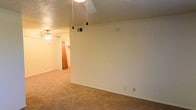 This single-story space presents a practical and straightforward layout designed for comfortable everyday living. The main living area features wall-to-wall beige carpeting that extends throughout, creating a warm and cohesive feel underfoot. A textured popcorn ceiling adds dimension overhead, complemented by a ceiling fan that provides both air circulation and ambient lighting.

Pendant light fixtures hang from the ceiling in the primary living space, offering a simple yet functional lighting solution. The walls are finished in a neutral off-white tone, providing a clean, versatile backdrop adaptable to various décor styles. A visible HVAC vent in the ceiling ensures climate control throughout the space.

An open passageway connects the main living area to an adjacent room, which also features a ceiling fan and appears to lead toward additional spaces, including what may be a kitchen or hallway. The layout promotes a natural flow between areas, making the space feel connected and accessible. Overall, this space offers a no-frills, functional environment with essential amenities in place, suitable for those seeking a straightforward and easy-to-maintain living arrangement.