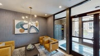Located at 44 Veterans Memorial Dr in Somerville, New Jersey, this contemporary building lobby makes a strong first impression with its thoughtfully curated design and upscale finishes. The polished marble-look tile flooring runs throughout, providing a clean and sophisticated foundation that ties the space together seamlessly.

At the heart of the lobby is a warm, light wood reception desk accented by striking black cage-style pendant lights that hang overhead. Behind the desk, a sleek open shelving unit with dark metal framing houses resident mailboxes, adding both function and visual interest. A dedicated package locker system, visible through floor-to-ceiling glass doors, ensures secure and convenient parcel management for residents.

A welcoming lounge area features a curated seating arrangement of rich amber-toned leather armchairs surrounding a low rectangular coffee table, all anchored by a patterned area rug. Overhead, a sculptural brass-and-glass globe chandelier adds a bold, modern focal point. The walls are finished in a textured gray wallcovering, and a pair of contemporary abstract artworks contribute color and personality to the space.

A stainless steel elevator with brushed metallic doors provides vertical access throughout the building. Large floor-to-ceiling windows allow natural light to fill the entry zone, creating a bright and inviting atmosphere. The overall design balances modern aesthetics with practical amenities, offering residents a polished and functional arrival experience.