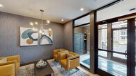 Located at 44 Veterans Memorial Dr in Somerville, New Jersey, this contemporary building lobby makes a strong first impression with its thoughtfully curated design and upscale finishes. The polished marble-look tile flooring runs throughout, providing a clean and sophisticated foundation that ties the space together seamlessly.

At the heart of the lobby is a warm, light wood reception desk accented by striking black cage-style pendant lights that hang overhead. Behind the desk, a sleek open shelving unit with dark metal framing houses resident mailboxes, adding both function and visual interest. A dedicated package locker system, visible through floor-to-ceiling glass doors, ensures secure and convenient parcel management for residents.

A welcoming lounge area features a curated seating arrangement of rich amber-toned leather armchairs surrounding a low rectangular coffee table, all anchored by a patterned area rug. Overhead, a sculptural brass-and-glass globe chandelier adds a bold, modern focal point. The walls are finished in a textured gray wallcovering, and a pair of contemporary abstract artworks contribute color and personality to the space.

A stainless steel elevator with brushed metallic doors provides vertical access throughout the building. Large floor-to-ceiling windows allow natural light to fill the entry zone, creating a bright and inviting atmosphere. The overall design balances modern aesthetics with practical amenities, offering residents a polished and functional arrival experience.