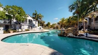 This single-floor property is set within a well-maintained residential community that exudes a distinct tropical, resort-like atmosphere. The standout feature of the outdoor space is a generously sized swimming pool with clear, turquoise water, complemented by a decorative rock water feature that adds a naturalistic focal point to the setting. The pool deck offers ample space for lounging, with seating areas positioned to take full advantage of the sunny, open-air environment.

The surrounding landscape is richly appointed with towering palm trees, mature tropical foliage, and carefully tended greenery that frames the community buildings and creates a lush, verdant backdrop. A charming gazebo structure is visible in the distance, adding architectural character to the grounds.

The community buildings present a clean, white exterior aesthetic with multi-level balconies, large windows, and classic architectural detailing. A spacious paved courtyard area with additional lounge seating provides a secondary outdoor gathering space, shaded by a canopy of mature trees. The overall ambiance of the outdoor areas is bright, airy, and reminiscent of a Florida resort community, bathed in abundant natural sunlight beneath clear blue skies. The combination of manicured landscaping, resort amenities, and elegant architecture creates a relaxed yet refined living environment.