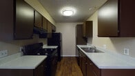 This single-floor residential unit offers a practical and cohesive layout designed for comfortable everyday living. The galley-style kitchen is a standout feature, lined on both sides with rich, dark espresso cabinetry that provides ample upper and lower storage. Light-toned countertops contrast the dark cabinetry, and the kitchen is equipped with a black gas range, over-range ventilation hood, refrigerator, and a double-basin stainless steel sink. Wood-style plank flooring runs throughout the kitchen, lending warmth and visual continuity. The open living and dining areas continue this flooring, creating a spacious, connected feel with neutral warm-beige walls and a textured ceiling. A utility closet with built-in ventilation is conveniently integrated into the living space. The bedroom features plush carpeting underfoot and benefits from natural light through a window with blinds, complemented by a ceiling fan for added comfort. The bathroom is finished with dark wood cabinetry beneath a single-basin vanity, a well-lit mirror with a multi-bulb fixture above, and wood-style flooring that ties the space together. Throughout the unit, the consistent use of dark cabinetry, warm neutral tones, and wood-style flooring creates a unified and inviting atmosphere.