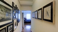 This single-floor space is defined by a well-appointed hallway that serves as both a transitional passage and a curated art gallery. The corridor's walls are lined with an eclectic collection of framed artwork and photographs — black-framed pieces on the left and a mix of sketch-style prints on the right — creating a visually engaging walkway that immediately establishes a creative, personalized atmosphere. The hallway features classic crown molding along the ceiling, adding an architectural detail that lends a sense of refinement to the space. Warm hardwood flooring runs throughout, catching natural light that filters in from the room at the end of the corridor. That room, visible through a doorway framed by a white door, appears to be a bedroom, illuminated by a large window that floods the space with bright, natural light. The bedroom features a made bed with rich purple and blue accent pillows, complementing the cool, neutral tones of the walls. The overall ambiance of the space balances a homey warmth with an artistic sensibility, making purposeful use of wall space to showcase personality and style throughout.