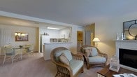 Located at 1000 Bally Bunion Drive in Egg Harbor, NJ, this single-floor residence offers a well-designed, open-concept layout that maximizes functionality and comfort. The living area is warm and inviting, featuring plush carpeting and ample space for relaxed seating arrangements, as evidenced by the classic rattan armchairs with light blue cushions that add a coastal, resort-like charm to the room. A pass-through counter connects the living space seamlessly to the kitchen, which is outfitted with white cabinetry, modern appliances including a full-size refrigerator and built-in microwave, and a clean, bright aesthetic enhanced by under-cabinet lighting. The open sightlines between the kitchen and living room create an airy, connected feel throughout the main living space. Neutral tones, including soft beige walls and light carpeting, lend a calm and cohesive ambiance to the interior. Additional rooms are accessible through connecting hallways, offering privacy and separation from the main living area. The overall design balances practicality with a relaxed, comfortable atmosphere, making this a well-appointed single-level residence in Egg Harbor, NJ.