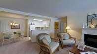 Located at 1000 Bally Bunion Drive in Egg Harbor, NJ, this single-floor residence offers a well-designed, open-concept layout that maximizes functionality and comfort. The living area is warm and inviting, featuring plush carpeting and ample space for relaxed seating arrangements, as evidenced by the classic rattan armchairs with light blue cushions that add a coastal, resort-like charm to the room. A pass-through counter connects the living space seamlessly to the kitchen, which is outfitted with white cabinetry, modern appliances including a full-size refrigerator and built-in microwave, and a clean, bright aesthetic enhanced by under-cabinet lighting. The open sightlines between the kitchen and living room create an airy, connected feel throughout the main living space. Neutral tones, including soft beige walls and light carpeting, lend a calm and cohesive ambiance to the interior. Additional rooms are accessible through connecting hallways, offering privacy and separation from the main living area. The overall design balances practicality with a relaxed, comfortable atmosphere, making this a well-appointed single-level residence in Egg Harbor, NJ.