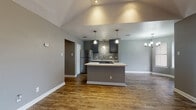 This single-story home showcases a thoughtfully designed open-concept floor plan that seamlessly connects the main living areas. The living room is highlighted by vaulted ceilings with recessed lighting and a ceiling fan, along with a sliding glass door fitted with vertical blinds that invites natural light and provides outdoor access. Rich hardwood-style flooring flows consistently throughout the entire home, lending warmth and visual continuity to each space.

The kitchen serves as a central hub of the home, featuring a substantial island with a sleek quartz-style countertop and an integrated cooktop. Dark gray cabinetry pairs with a mosaic tile backsplash, stainless steel appliances — including a refrigerator and dishwasher — and pendant lighting overhead. A separate dining area is defined by an elegant chandelier.

The bedrooms are generously proportioned, finished in neutral gray tones with large windows offering natural light. One bedroom includes direct access to a full bathroom equipped with a gray vanity, framed mirror, and a soaking tub surrounded by large-format marble-look tile with brushed bronze fixtures. The overall aesthetic is clean, modern, and move-in ready.