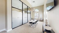 Located at 1400 Village Blvd in West Palm Beach, FL, this thoughtfully designed office space offers a refined, contemporary environment ideal for professional use. The single-floor layout encompasses a well-appointed office that serves multiple functions, combining a central conference area, dedicated workstations, and a comfortable lounge seating arrangement.

The conference area is anchored by a sleek white rectangular table surrounded by modern cantilever chairs with upholstered seats. Overhead, two statement crystal bead chandeliers add an elegant touch, drawing the eye upward to the impressively tall ceilings. A striking floor-to-ceiling glass partition wall with black framing and frosted lower panels divides the interior, allowing light to flow throughout while maintaining a sense of privacy.

Built-in workstations line the far wall beneath a decorative geometric wallcovering accented with circular wall art, creating a focused work area. Natural light pours in through multiple windows fitted with horizontal blinds, while an arched upper window frames views of lush tropical greenery outside. A wall-mounted flat-screen television and a stylish console table with cane detailing add functional elegance to the space.

Direct outdoor access through a glass door opens to an exterior terrace, enhancing the bright, airy ambiance. The neutral carpeted flooring, warm beige walls, and curated décor throughout give this office a polished, welcoming atmosphere.