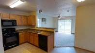 This single-floor space offers a functional and open layout that seamlessly connects the kitchen and living area. The kitchen is well-appointed with warm honey-toned wood cabinetry providing ample storage, dark granite-style countertops, and a full suite of black appliances including a range, built-in microwave, and dishwasher. A double-basin sink is positioned along the peninsula counter, which acts as a natural divider between the kitchen and the adjoining living space. Tile flooring runs throughout the kitchen, while plush carpeting extends across the living area, adding a sense of comfort and warmth. The living room benefits from abundant natural light streaming through large sliding glass doors fitted with vertical blinds, which open to an outdoor balcony or patio, providing views of the surrounding greenery. A ceiling fan is centrally located in the living area, enhancing air circulation. Additional lighting in the kitchen is provided by track-style spotlights and a fluorescent fixture. A wall-mounted air conditioning unit is also present, ensuring year-round comfort. The textured ceilings add a classic finishing touch throughout the space.