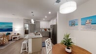 This single-floor residence offers a thoughtfully designed open-concept layout that seamlessly connects the kitchen, dining, and living areas. The kitchen is a standout feature, showcasing two-tone cabinetry with white upper and gray lower cabinets, granite countertops, stainless steel appliances, and a large center island with seating — illuminated by elegant pendant lighting. A built-in wine rack adds a refined touch, while light wood-style flooring flows throughout the main living areas.

The living room is bright and welcoming, with large windows dressed in sheer curtains that invite natural light, complemented by a ceiling fan for added comfort. A glass-top coffee table and neutral furnishings contribute to the airy, coastal-inspired ambiance.

The bedrooms feature plush carpet, ceiling fans, and generous natural light. The primary bedroom is accompanied by an en-suite bathroom with dual granite vanities, a glass-enclosed walk-in shower, and direct access to a spacious walk-in closet. A second bathroom offers a tiled shower-tub combination and granite countertops. An in-unit laundry room with a full-size washer and dryer provides everyday convenience, completing this well-equipped and comfortable residence.