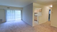 This single-floor residential unit offers a practical and comfortable layout designed for everyday living. The living area is carpeted and bathed in natural light, courtesy of a large sliding glass door fitted with vertical blinds that opens to an outdoor balcony, providing pleasant views of surrounding greenery. The open layout creates a sense of spaciousness, with neutral-toned walls that lend a clean and versatile aesthetic throughout.

Adjoining the living area is a well-appointed kitchen, visible through an open pass-through layout that maintains connectivity between the two spaces. The kitchen features light wood cabinetry, a double sink, and includes white appliances such as a refrigerator, offering ample storage and functional counter space. The kitchen flooring transitions from the carpeted living area to a smoother surface, clearly delineating the two zones.

The overall ambiance of the unit is bright, neutral, and move-in ready, with a straightforward design that emphasizes functionality. The combination of natural light from the balcony-facing glass door and the open connection between the living and kitchen areas makes the space feel welcoming and easy to personalize.