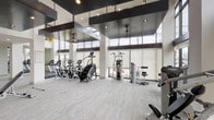This fitness center is designed with both functionality and aesthetics in mind, offering a well-appointed space for a full range of workout activities. The facility features soaring ceilings with bold, dark architectural beam accents that contrast sharply against the white walls and structural columns, creating a visually striking environment. Large floor-to-ceiling windows line the perimeter, flooding the space with natural light and providing open views to the outdoors, while roller shades offer adjustable light control.

The flooring throughout is a smooth, light gray stone-look surface that gives the space a clean, polished appearance. The cardio zone is well-equipped with StairMaster steppers, ellipticals, and treadmills arranged in an organized row along the windowed wall. The strength training area features a variety of cable machines, resistance stations, and functional training equipment. A free weight section with a dumbbell rack and adjustable bench rounds out the offerings.

A separate entry area serves as a welcoming transition zone, accented with vibrant wall art and glass-paneled doors. Mounted flat-screen televisions are positioned throughout for entertainment during workouts. Pendant lighting and ceiling fans add both style and comfort to this thoughtfully designed fitness facility.