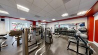 The Fitness Center at Arrive Stamford is a comprehensively equipped facility designed to support a full range of workout routines. The space features a drop-panel ceiling with recessed fluorescent lighting that ensures consistent, bright illumination throughout. A bold red accent band running along the upper perimeter of the walls adds a dynamic, energetic character to the otherwise neutral white interior.

The flooring throughout the center consists of durable rubber matting, providing traction and cushioning suited for both strength training and cardio activity. Large mirrors line multiple walls, visually expanding the space and allowing users to monitor their form during exercise.

The cardio zone is equipped with multiple treadmills and stationary bikes, each outfitted with individual console displays. Wall-mounted flat-screen televisions are positioned above the cardio equipment for entertainment during workouts. The strength training area includes a variety of cable and resistance machines, free-weight benches, and an extensive rack of dumbbells ranging across multiple weight increments. Additional equipment such as stability balls and a cable cross machine further diversify training options.

Natural light enters through windows and glass doors along one wall, which also provide views of an outdoor area. The overall layout is organized and spacious, allowing for comfortable movement between equipment stations.